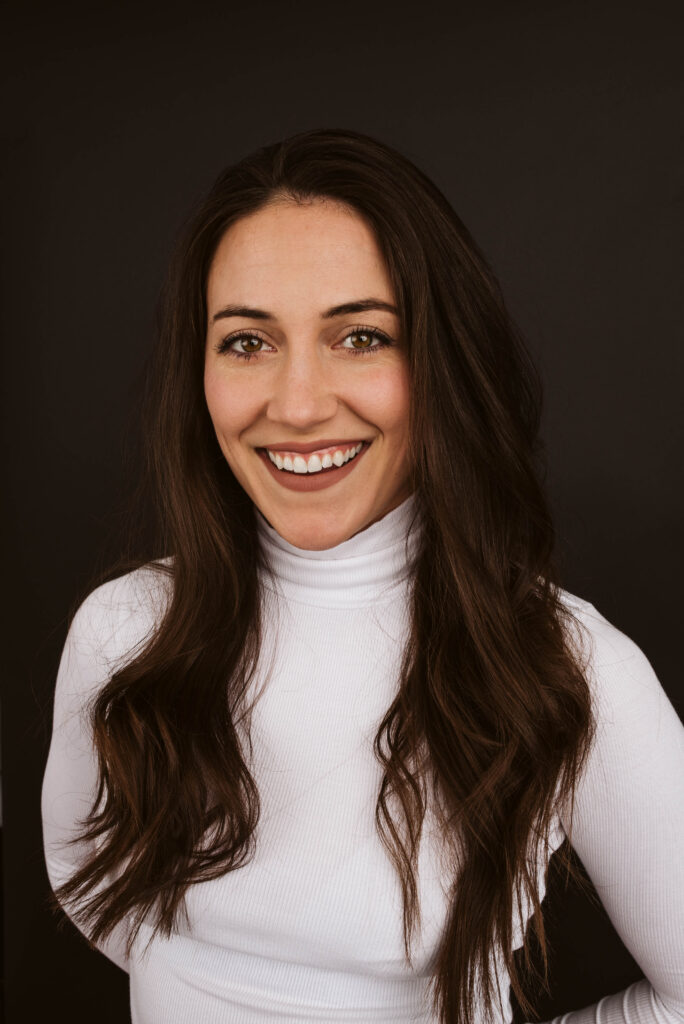 Smiling woman with long brown hair in a white turtleneck against a dark background.
