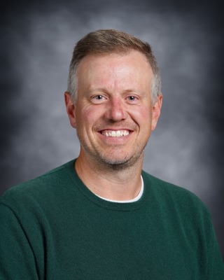 Head-and-shoulders portrait of a smiling man with short gray hair wearing a green crewneck sweater, against a gray studio backdrop.